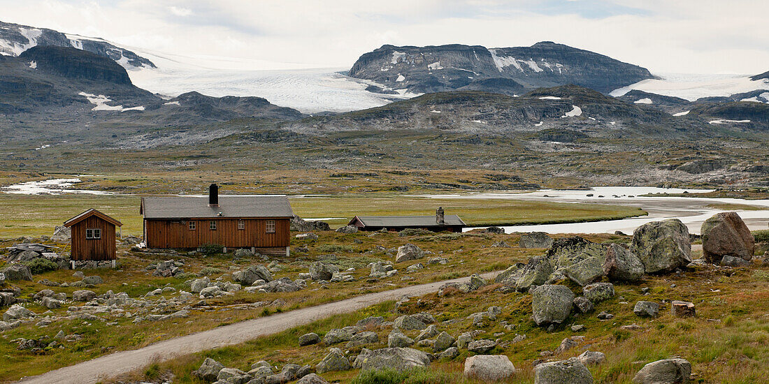 A Wooden House And Shed Stand Along A Fjord With A Dirt Road And Mountains In The Background; Highlands Norway