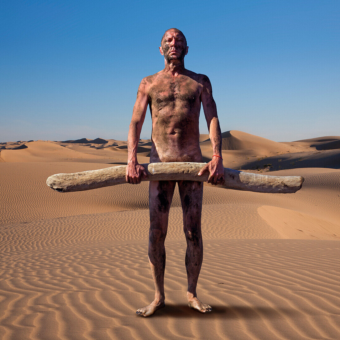 Nackter Mann mit Schmutz bedeckt, der Treibholz auf einer Sanddüne hält