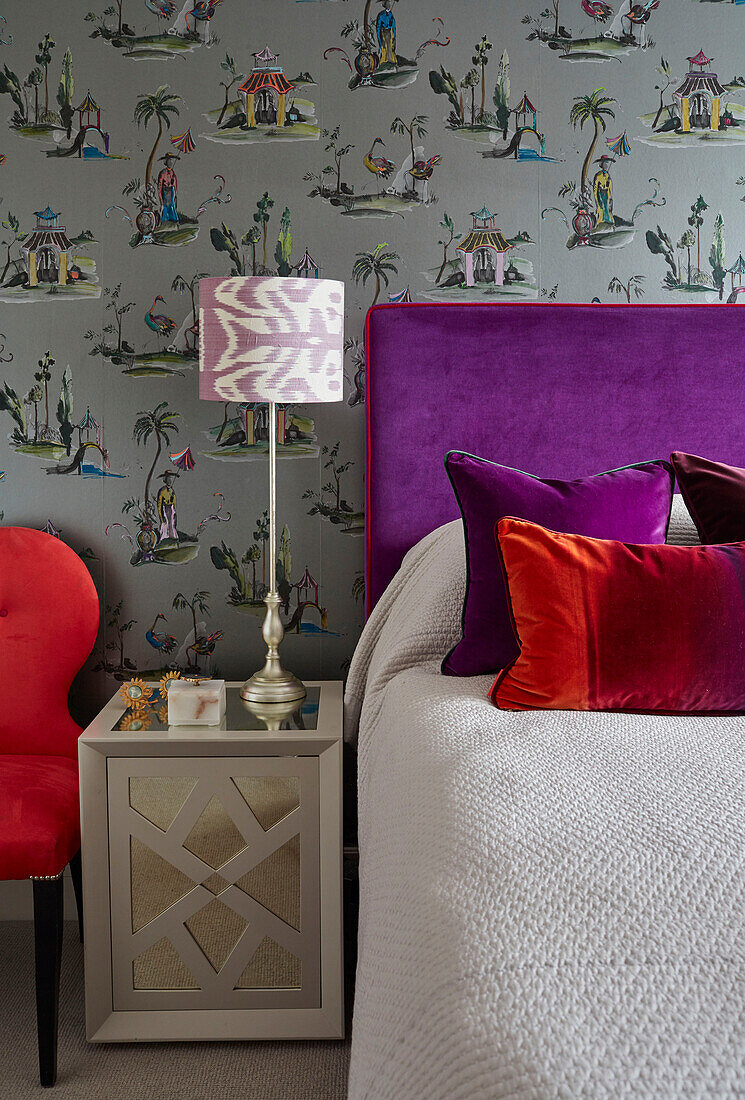 Purple headboard and Oriental wallpaper with red velvet pillows and chair in London apartment, UK