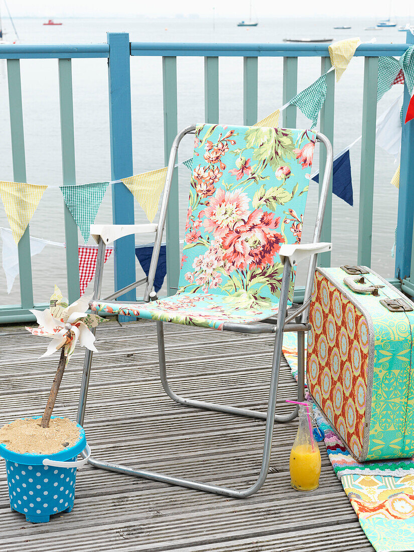 Floral patterned folding chair with vintage suitcase on coastal decking with bunting