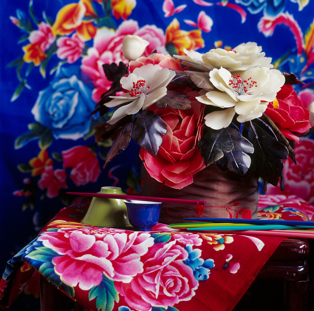 Still life of brightly coloured Chinese fabrics behind handmade Mexican wax flowers and other Chinese artifacts