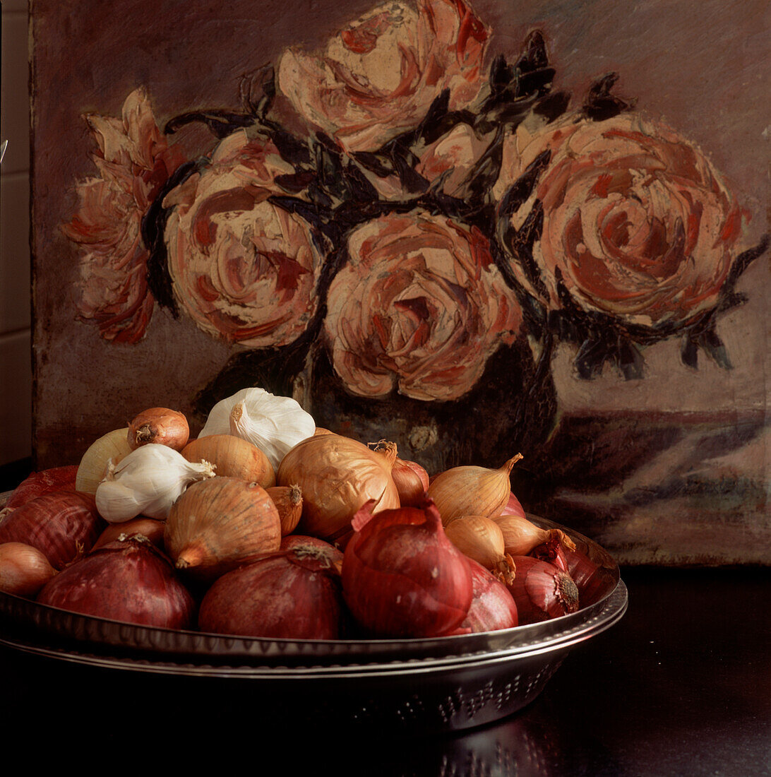 Stilleben mit Schale mit Zwiebeln und Knoblauch auf einem Tisch mit einem floralen Ölgemälde im Hintergrund