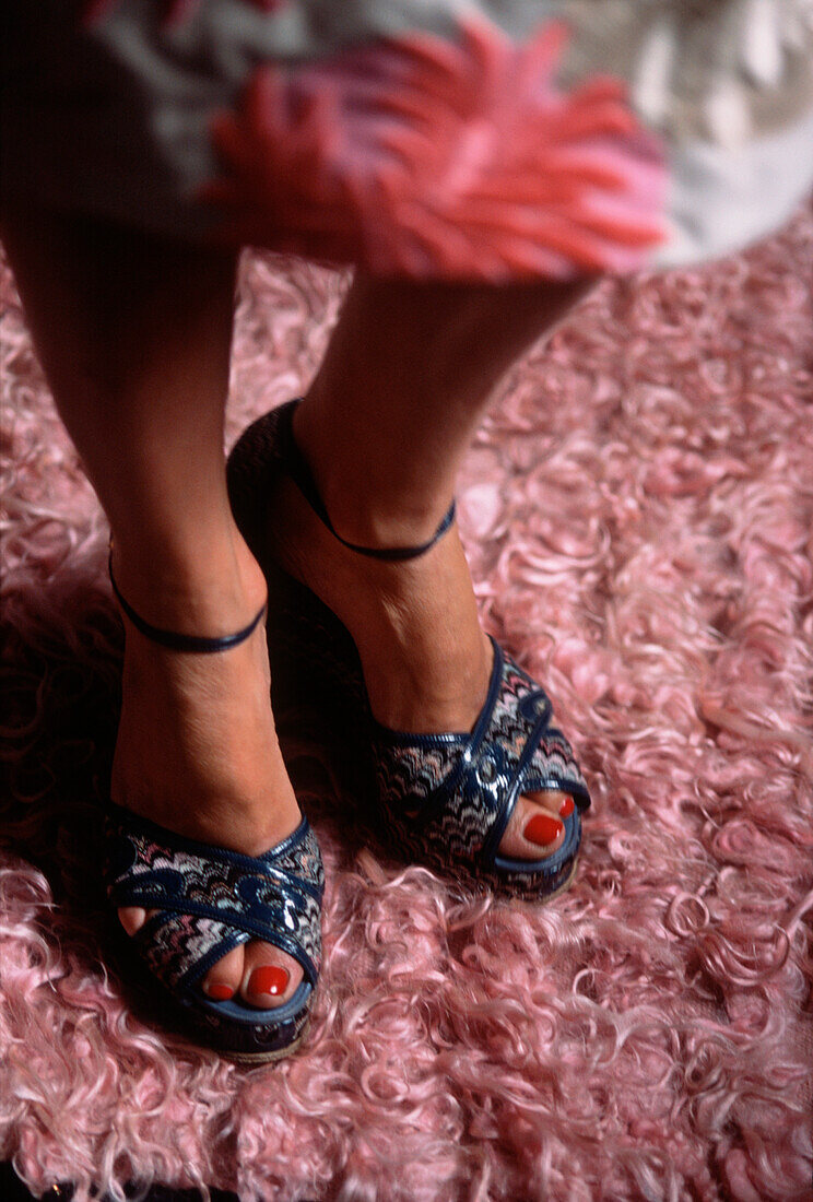 Detail of a woman's feet standing on a pink shag pile rug wearing open toed high heeled sandals with pink nail polish