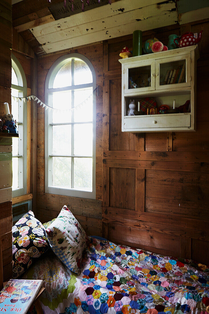 Sonnenbeschienenes Bogenfenster über einer Patchwork-Bettdecke im holzgetäfelten Baumhaus in Rye, East Sussex, England, UK