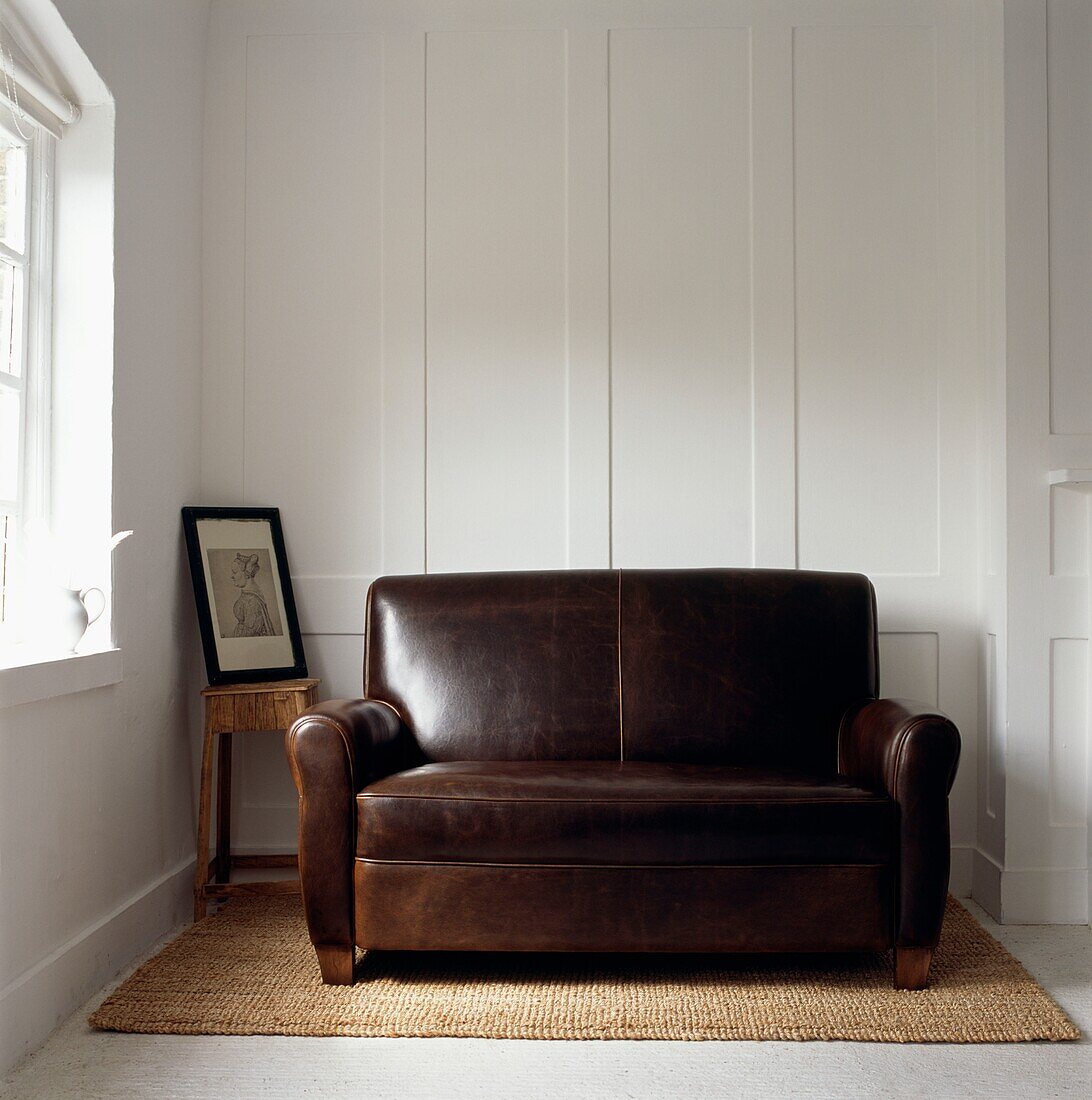 Zweisitzer-Sofa aus braunem Leder auf Seegras-Teppich mit weiß getäfelter Wand