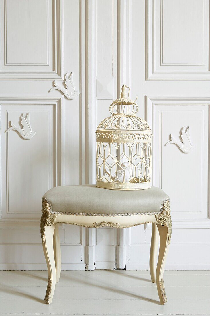 Upholstered stool with ornate bird cage an white dove decorations on a panelled wall