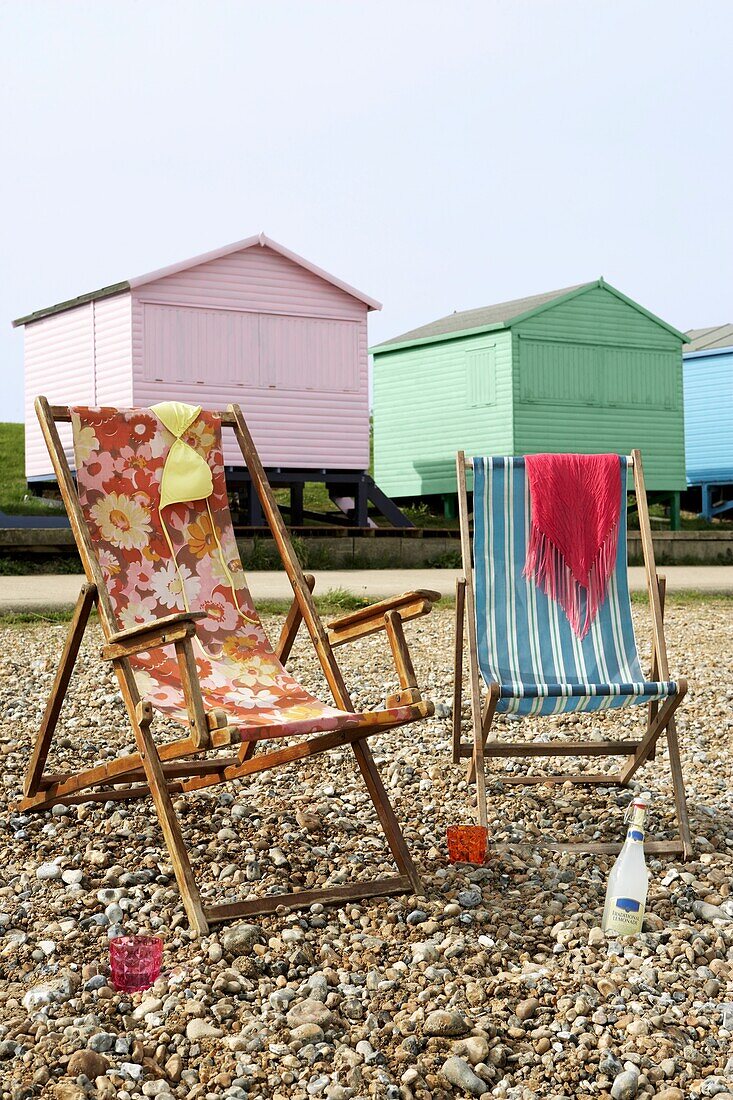 Two deck chairs on a pebble beach with colourful beach huts behind