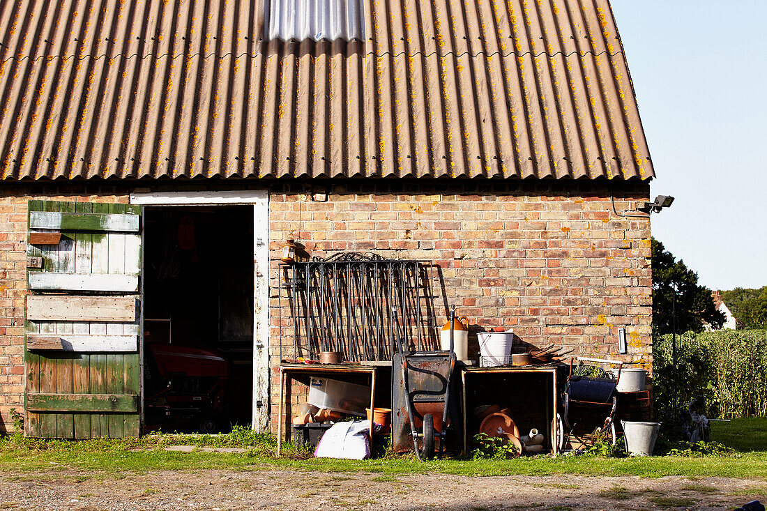 Wheelbarrow and gardening equipment,   brick barn exterior with corrugated roof,  Brabourne,  Kent,  UK