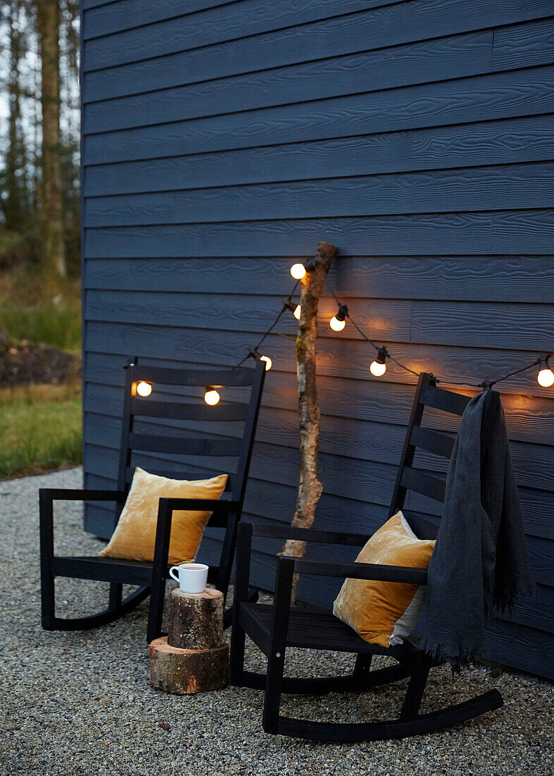 Ein Paar Schaukelstühle mit beleuchteten Lichtern an der holzverkleideten Außenfassade eines Neubaus in Devon (UK)