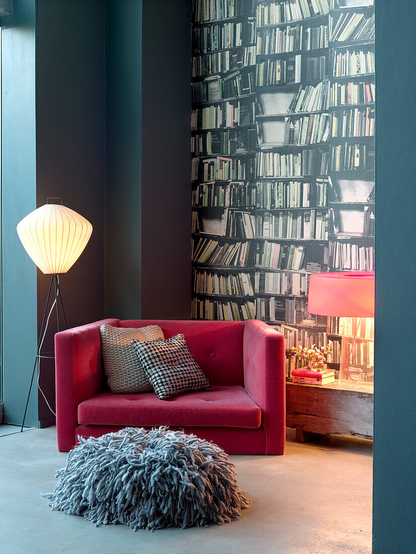 Rosafarbener Sessel an einer mit Büchertapete dekorierten Wand mit grauem Bodenkissen und Stehlampe