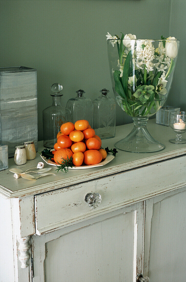 Flower arrangement in glass vase and pile of oranges on cupboard
