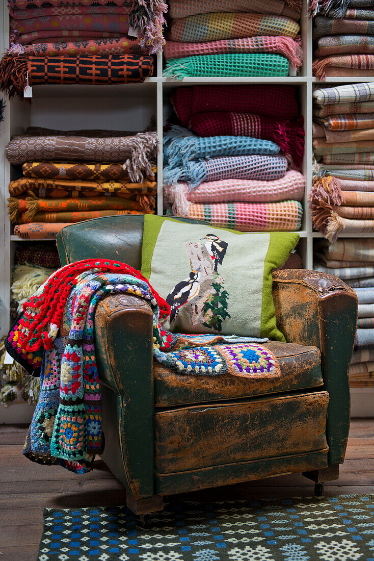 Crochet blankets and cushion on worn leather armchair with shelves of Welsh blankets in Tregaon shop interior Wales UK