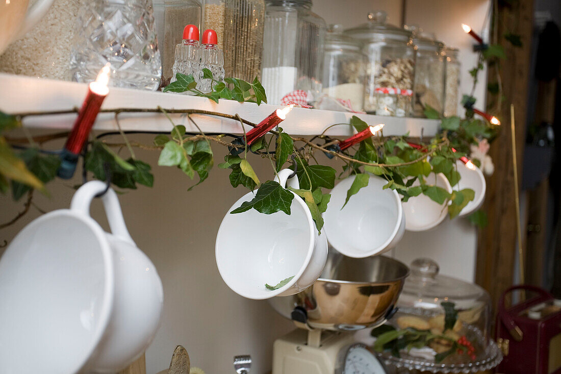 Teacups hang from shelving with fairylights in kitchen of Tenterden home, Kent, England, UK