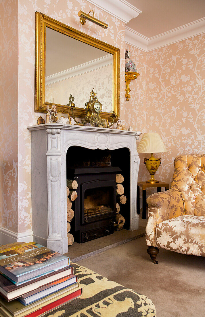 Upholstered armchair at fireplace with gilt framed mirror and wood burning stove in Kent home England UK