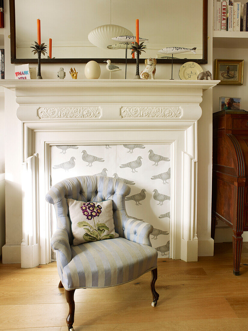 Upholstered armchair and cushion in front of fireplace with ornaments in London home England UK