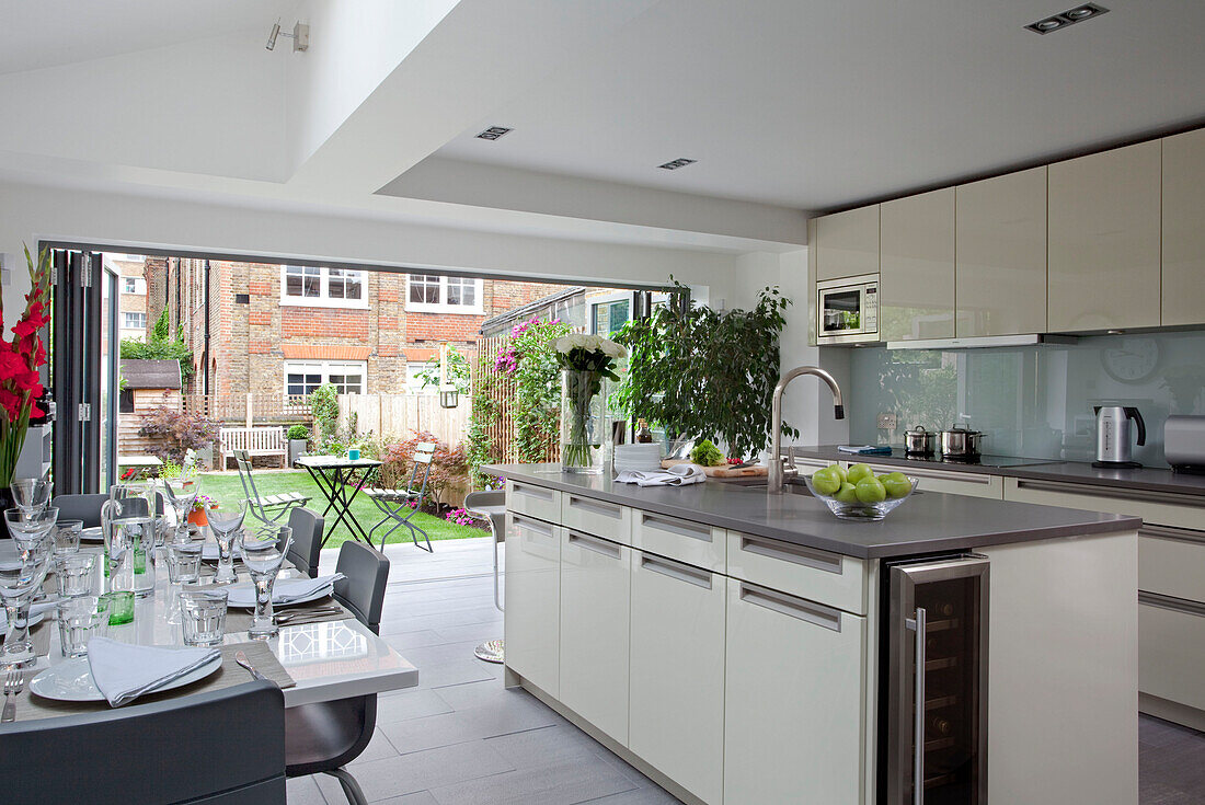 Weiße und graue offene Küche und Essbereich mit offenen Türen zum Garten in einem modernen Haus in London, England, UK