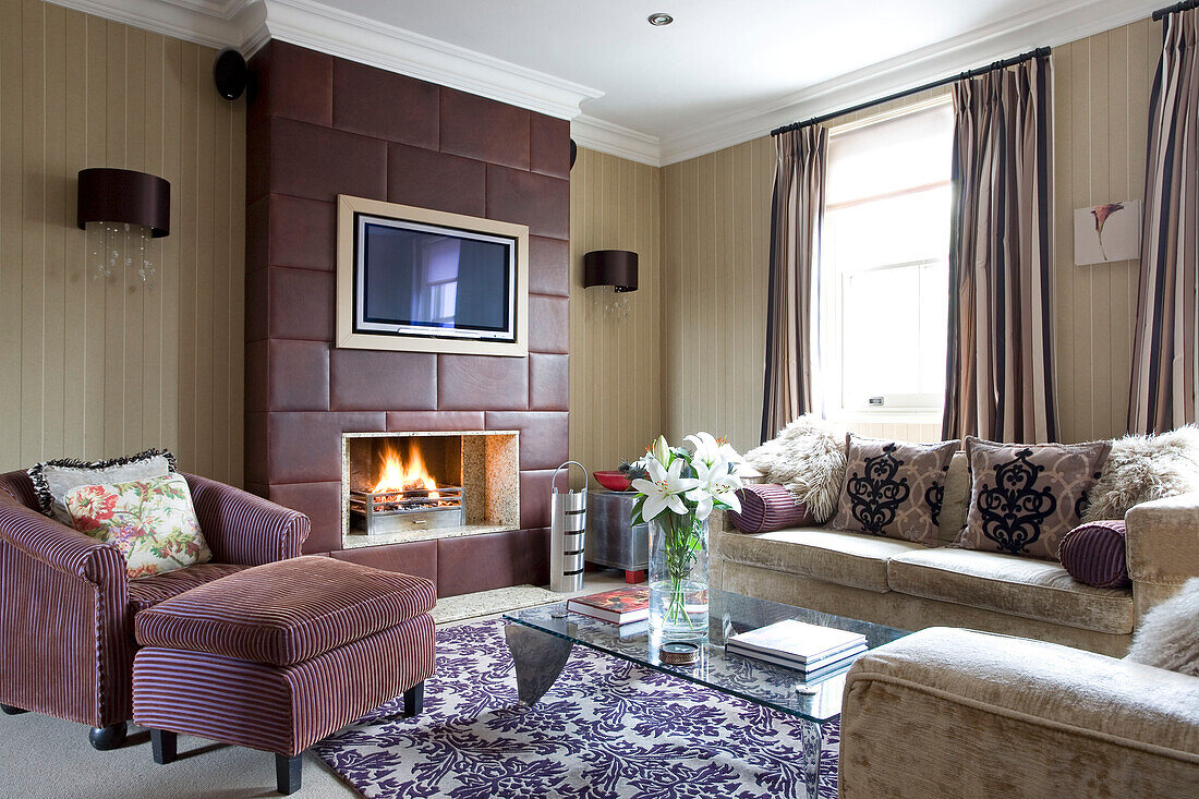 Armchair and footstool at fireside with brown leather chimney breast with plasma screen TV