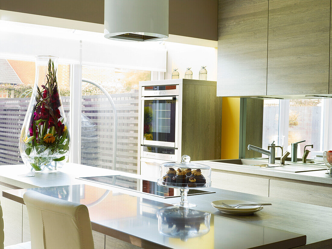 Large glass vase filled with flowers with cake stand and fitted electric hob on kitchen island of Manchester home, England, UK