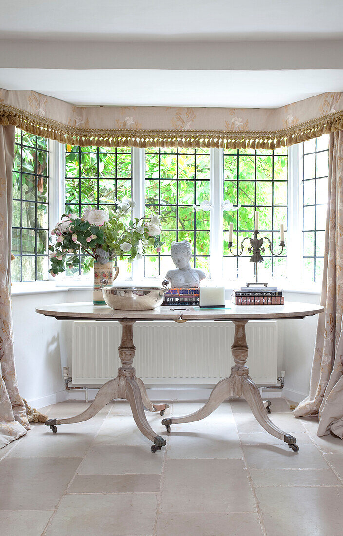 Ornamente und Bücher auf ovalem Tisch im Erker eines Bauernhauses in Sussex, UK