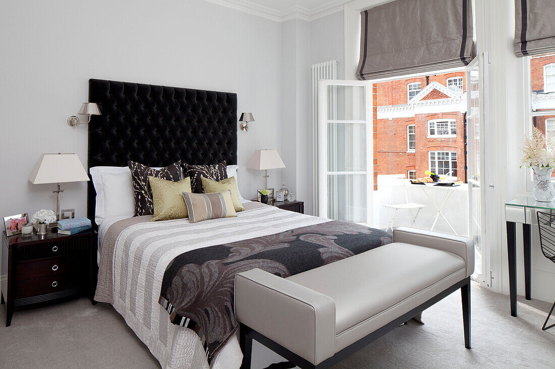 Bed with black buttoned headboard in room with open doors to balcony exterior, London apartment, UK