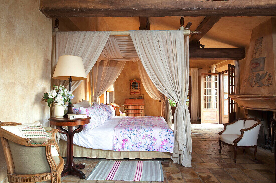 Four poster bed with cream fabric in stone floored bedroom of French holiday villa