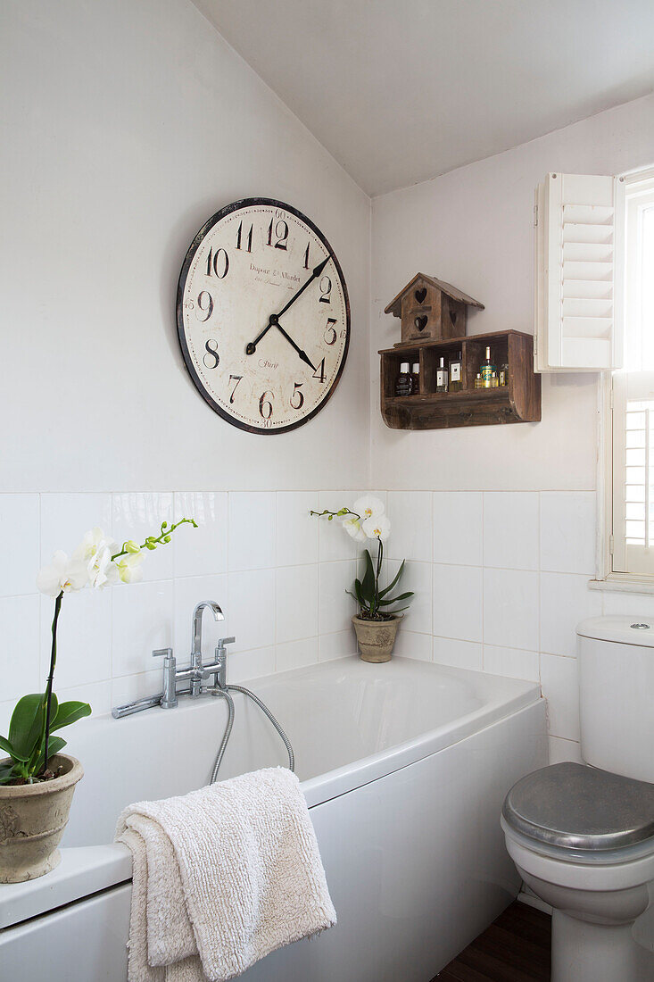 Large clock and shelving above orchids on white bath with mixer tap in Berkshire home,  England,  UK