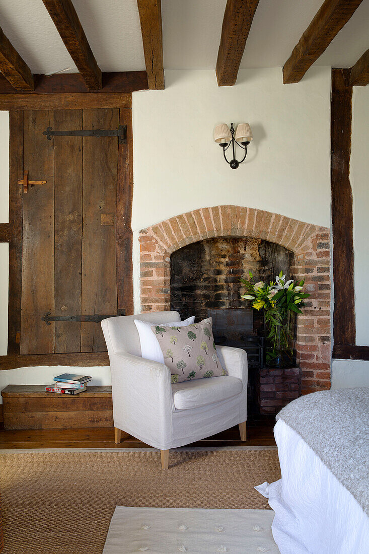 White armchair at brick fireplace in beamed living room of Sussex farmhouse   England   UK