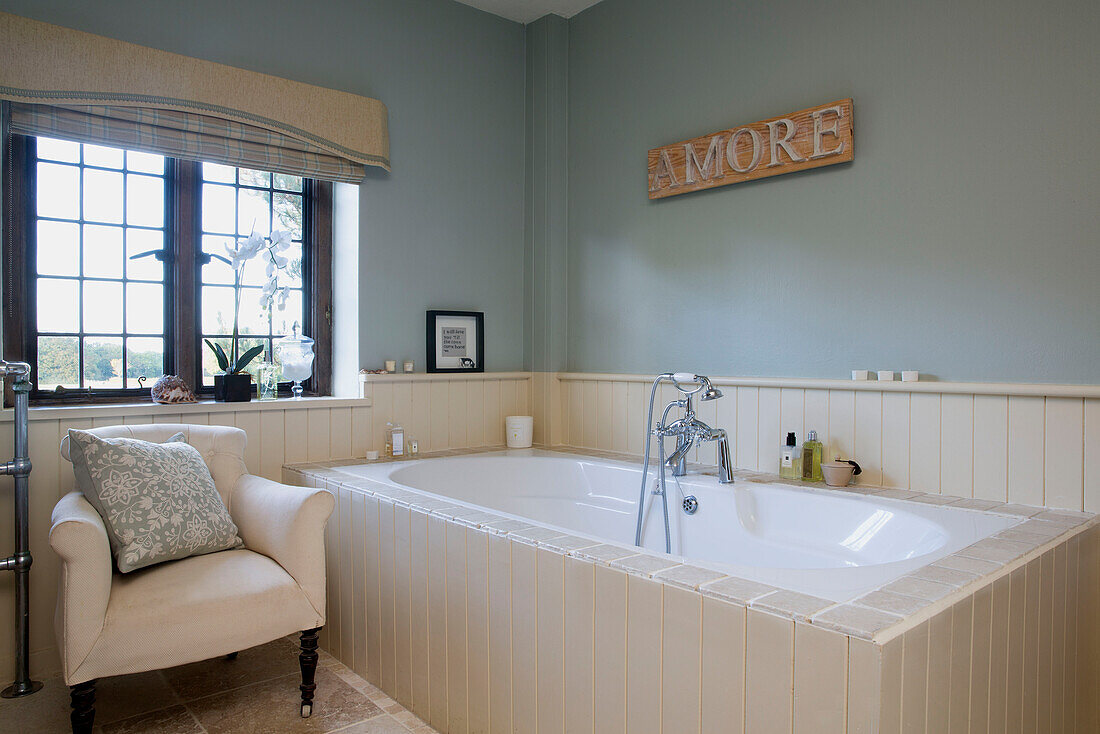 Cream armchair and bath with leaded window in Sussex farmhouse   England   UK