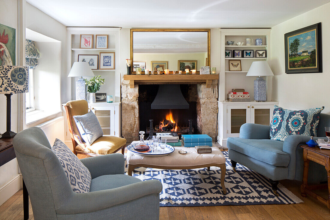 Light blue armchairs with gilt framed mirror above lit fire in Gloucestershire cottage UK