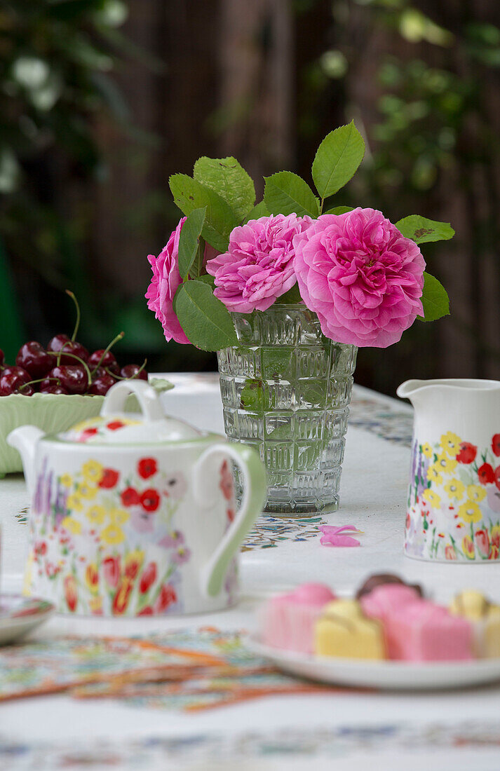 Rosa Schnittblumen mit geblümter Teekanne und Kirschen auf Gartentisch London UK