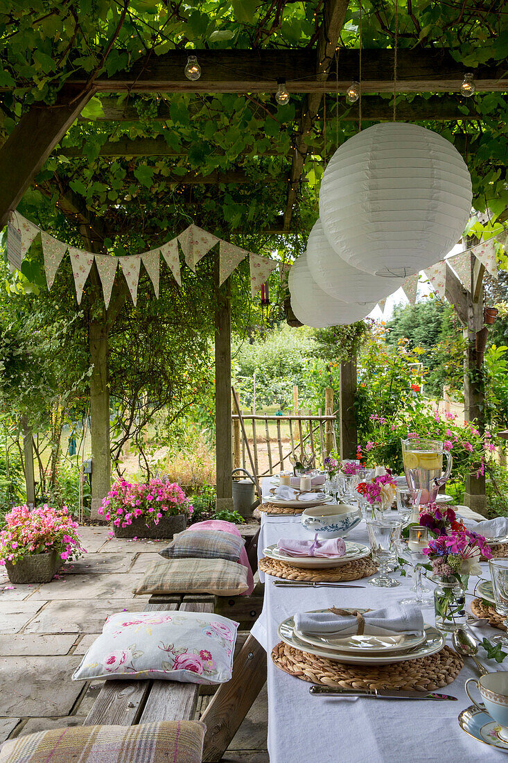 Paper shades hang from pergola above dining table with seat cushions and bunting in Alford garden Surrey UK