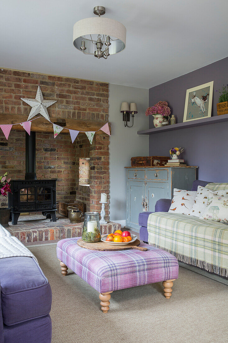 Checked pink ottoman and sofa with woodburner in exposed brick fireplace with bunting in detached 1950s house in Alford Surrey UK
