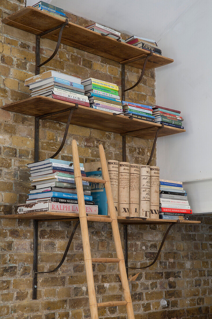 Wall mounted shelving and books with ladder on exposed brick wall in converted South London schoolhouse UK