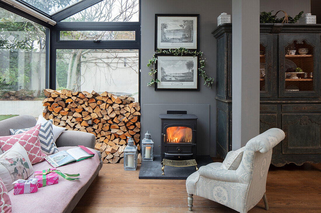 Sessel und Sofa mit Holzstapeln und Kaminfeuer im Wintergartenanbau einer Londoner Villa UK
