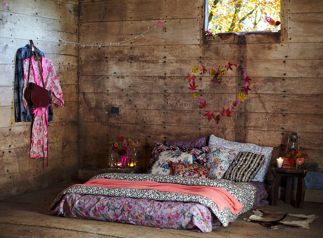 Verschiedene Blumenstoffe auf Matratze unter Blätterherz und Fenster in rustikaler Holzhütte Herbst UK