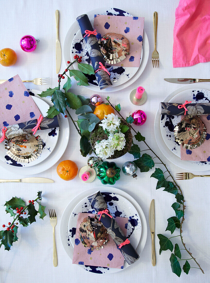 Overhead view of four places set on a white tablecloth for Christmas with crackers baubles holly and ivy