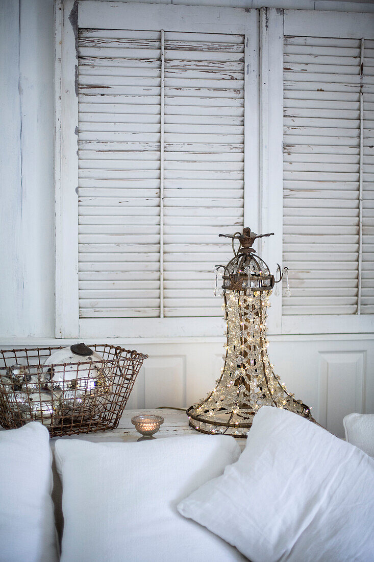 Scandi style living room with white cushions basket of silver baubles and chandelier with fairylights on and louvered shutters on the wall