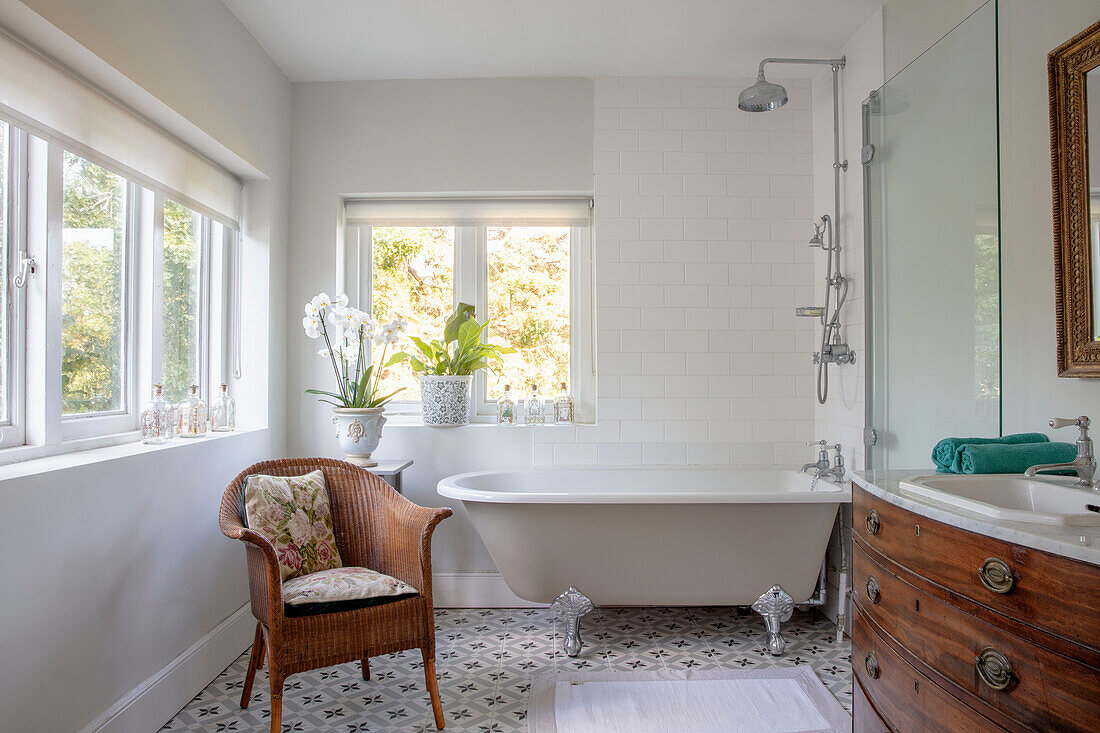 Freestanding bathtub with rain shower and rattan armchair in bathroom with patterned tiled floor
