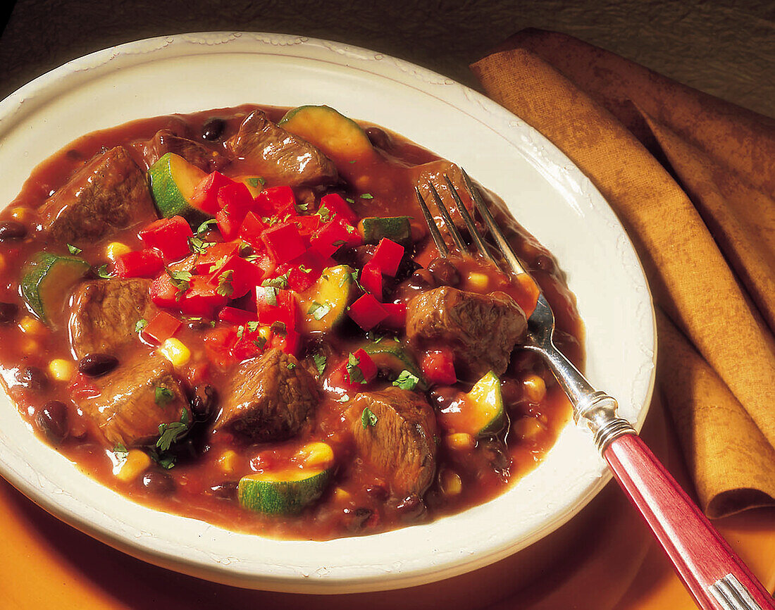 A bowl of Mexican style beef stew with a fork and golden napkin