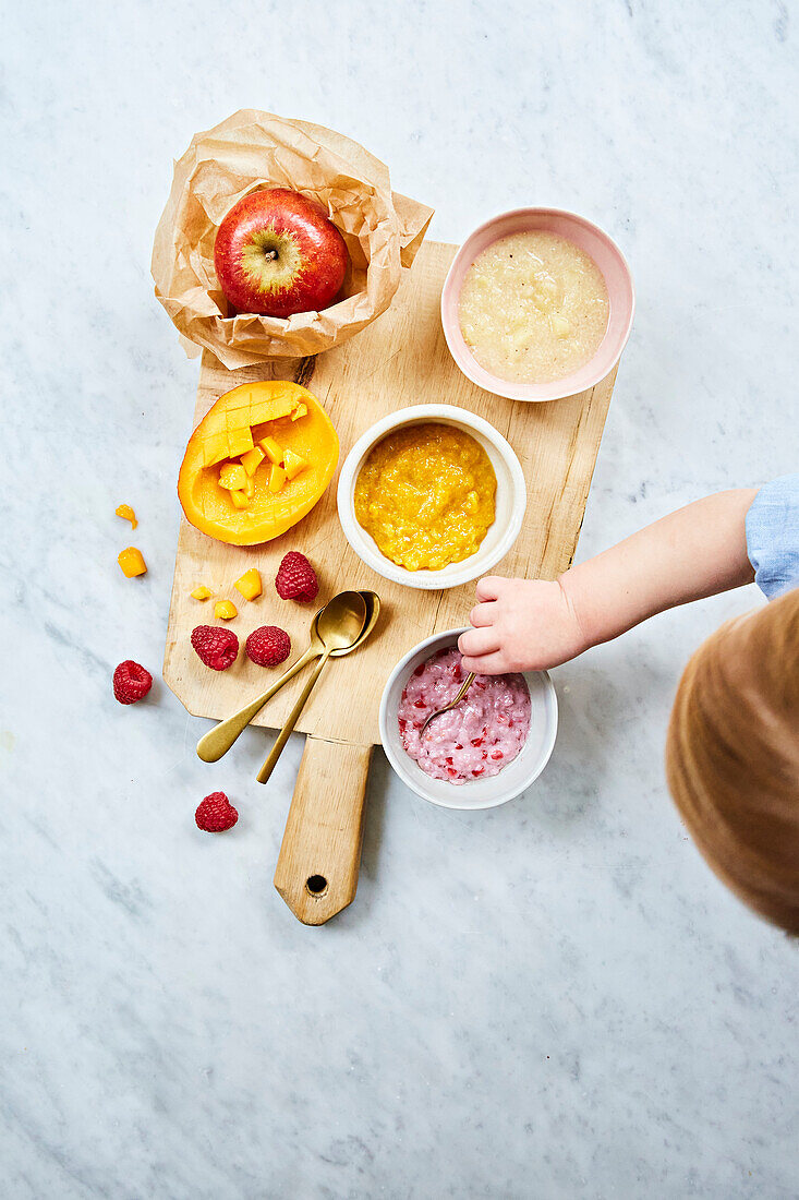 Children's fruit on a board with a toddler reaching in. Mango, raspberry, apple and banana