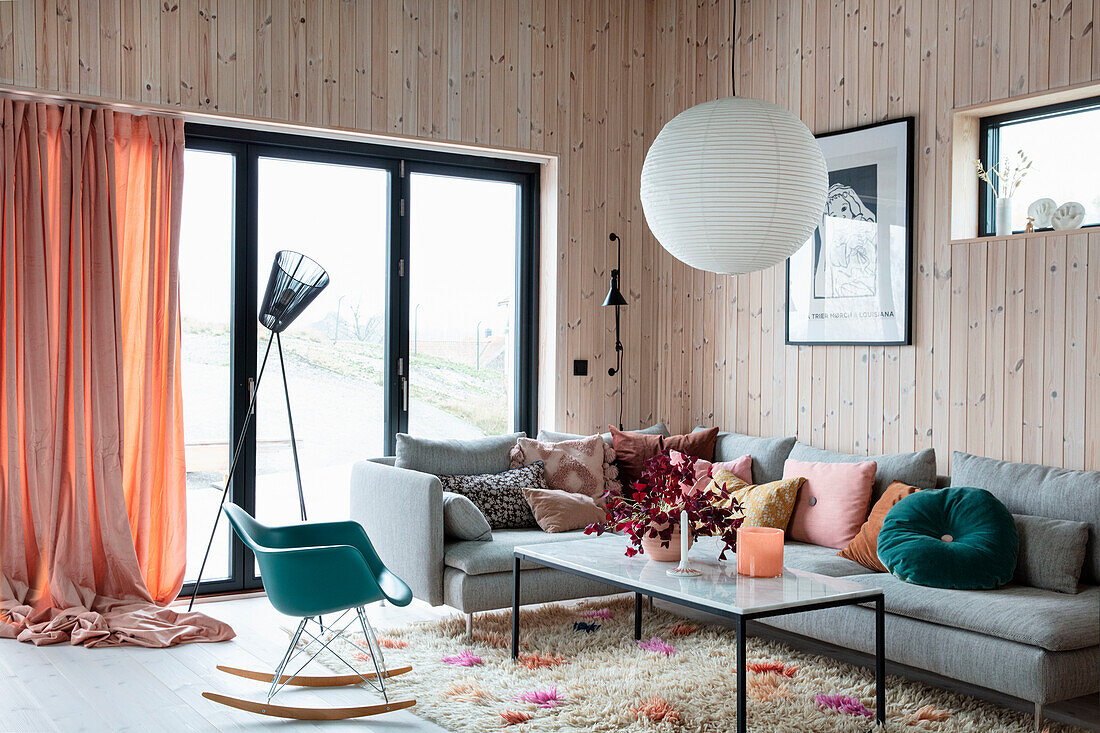 Wohnzimmer mit Holzwandverkleidung, Designerschaukelstuhl und bunten Kissen auf grauem Ecksofa