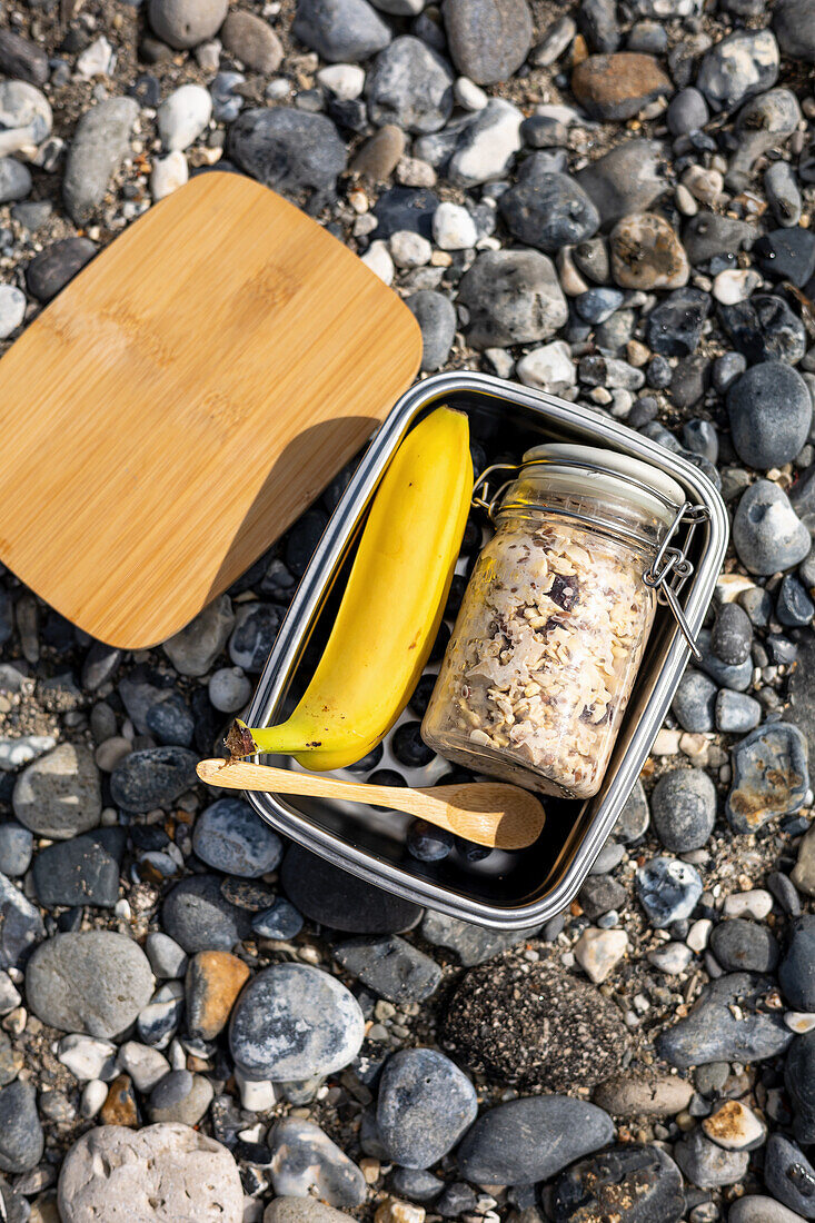 Overnight Oats mit Blaubeeren und eine Banane in Lunchbox auf Kieselsteinboden