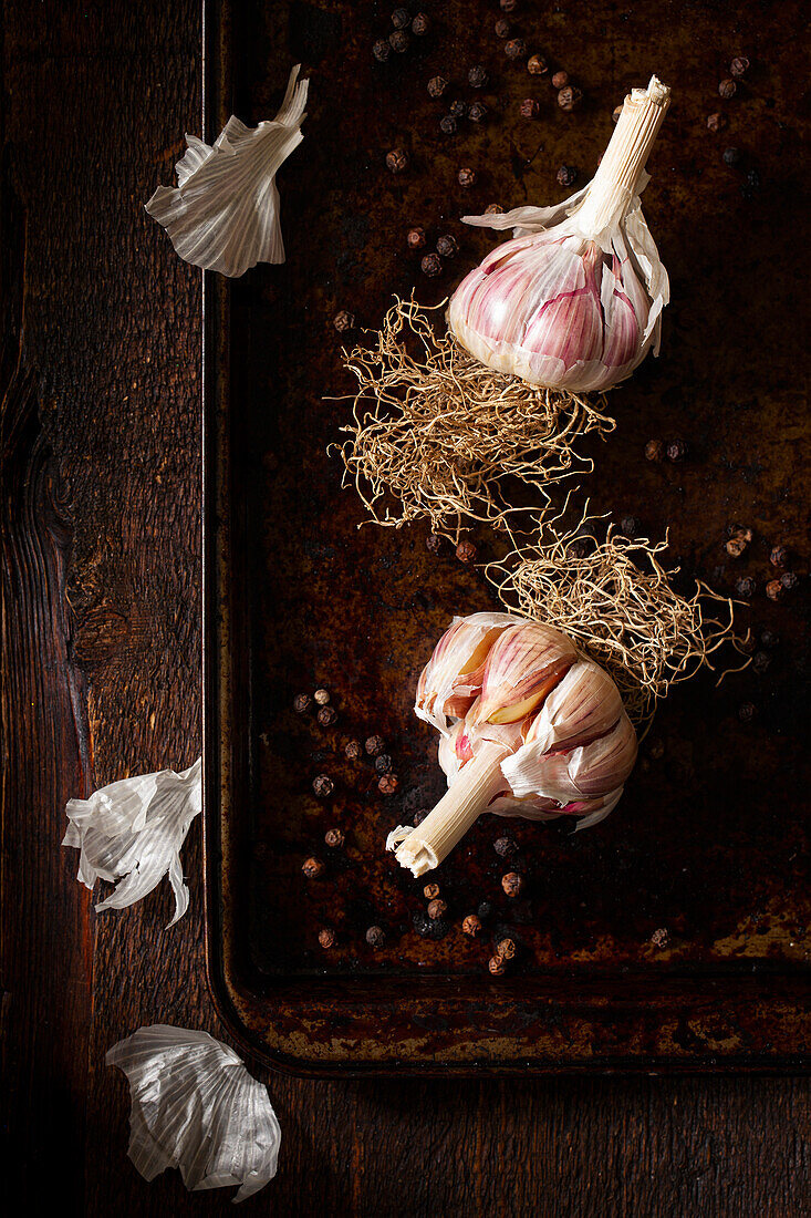 Two fresh garlic bulbs with roots still attached on a baking tray with black peppercorns