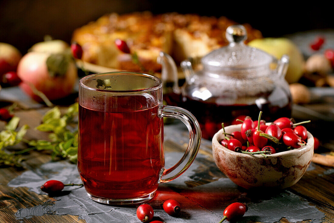 Rose hip berries herbal tea in glass mug on old wooden plank table with wild autumn berries and apple pie around