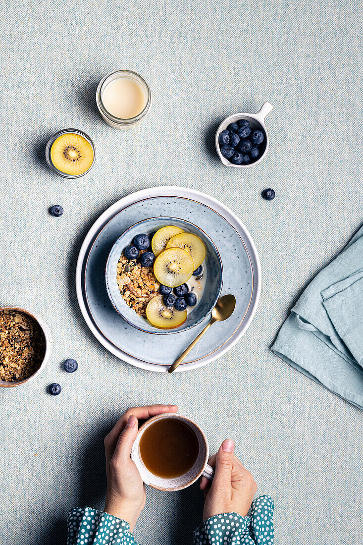 Warming hand with cup of hot drink at breakfast with bowl with granola and blueberries and kiwi slices
