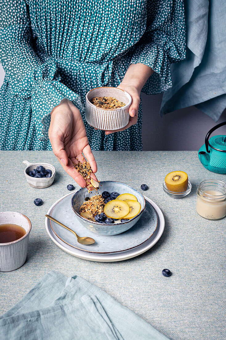 Crop anonymous female making healthy breakfast consisting of bowl of granola with blueberries and kiwi slices and cup of tea