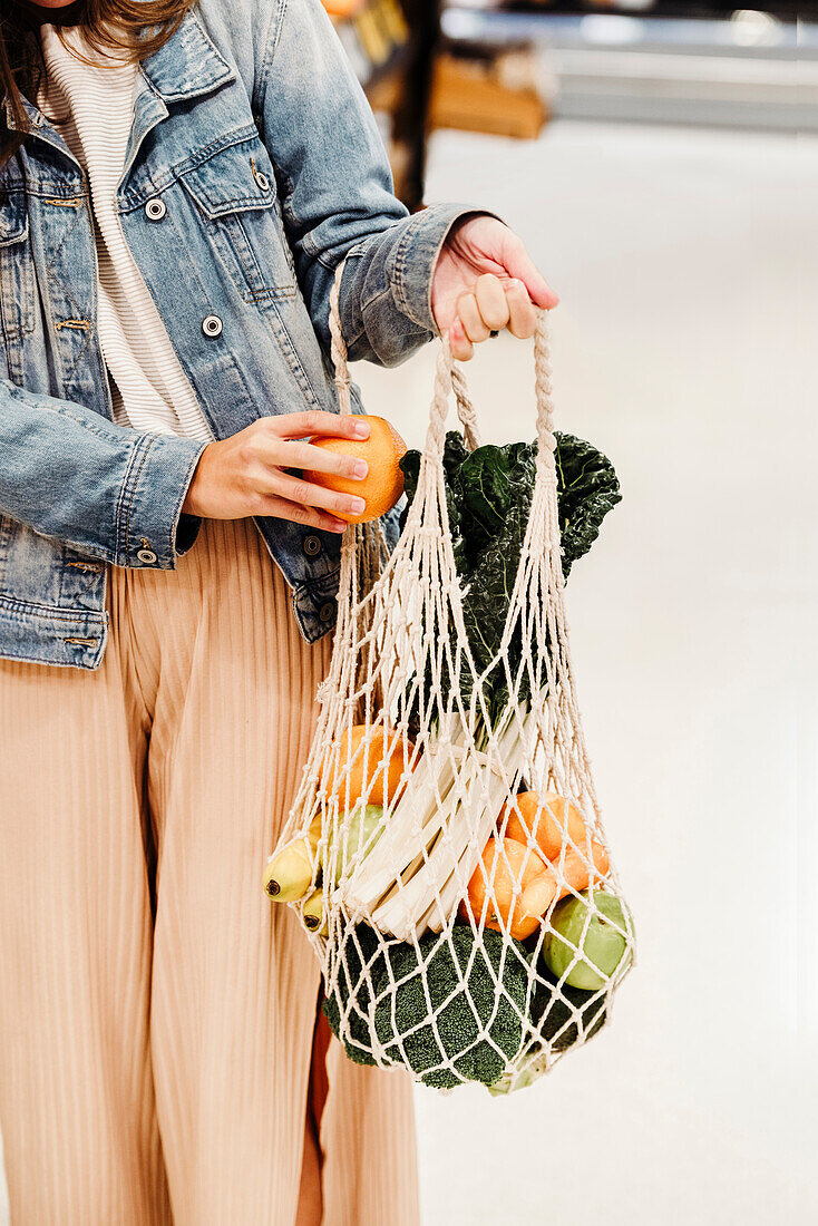 Cropped unrecognizable female standing with assorted fruits and vegetables in eco friendly mesh bag