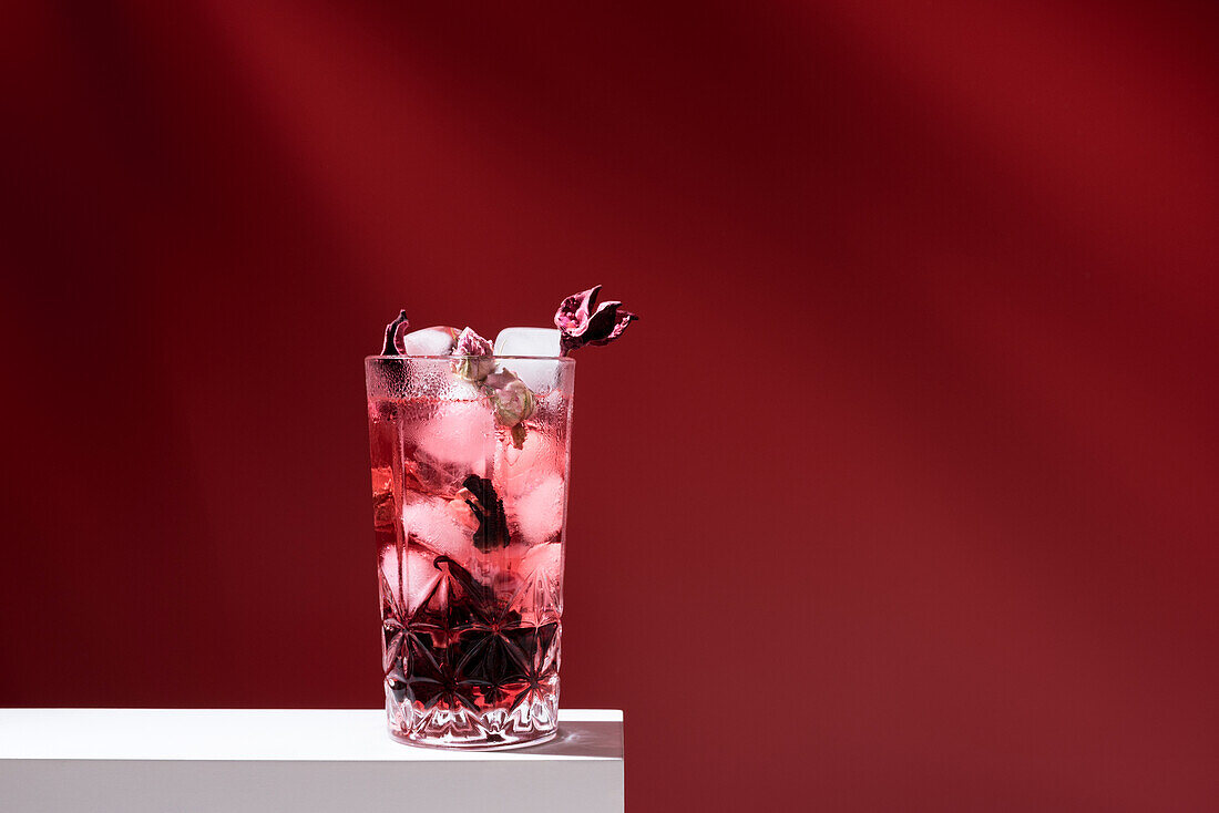 Glas mit rosafarbenem Gin Tonic und Eiswürfeln auf weißem Tisch vor rotem Hintergrund