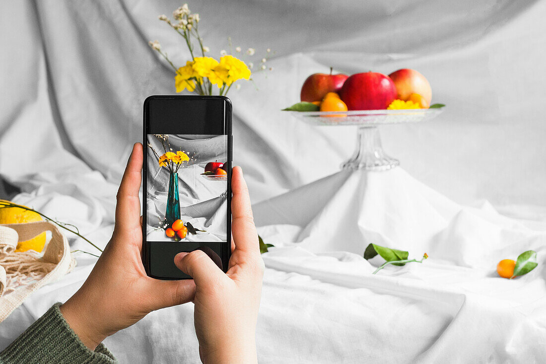 Crop anonymous person taking photo of fresh kumquats and blooming flower bouquet in vase on cellphone