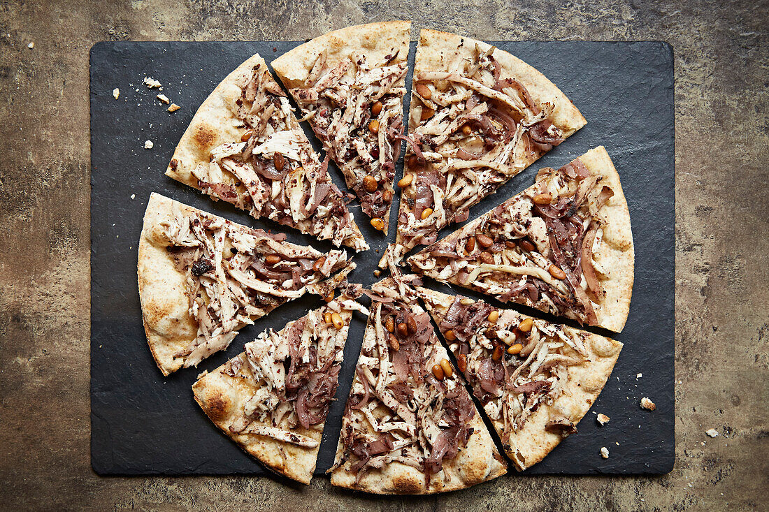 A pizza topped with chicken, pine nuts and red onions, sliced on a slate platter